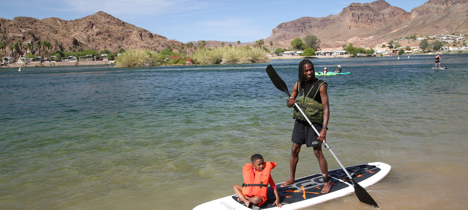 A man standing on a paddleboard with a child in a lifejacket sitting in front of him on the Colorado River