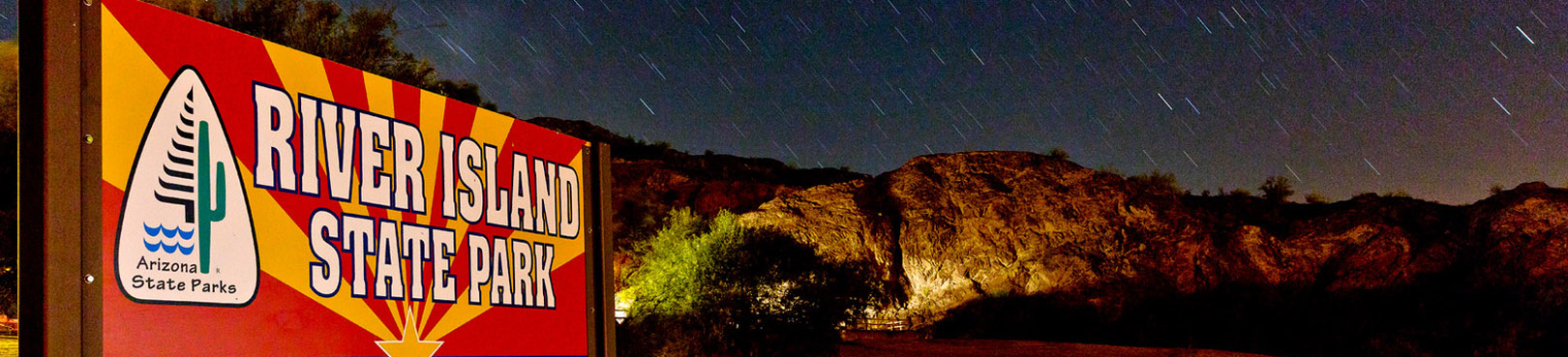 An illuminated River Island State Park sign and rugged desert mountains below a starry night.