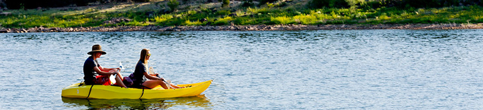 Two people in a bright yellow double kayak glide along the lake surface near the grass lined shore of Fool Hollow Lake.