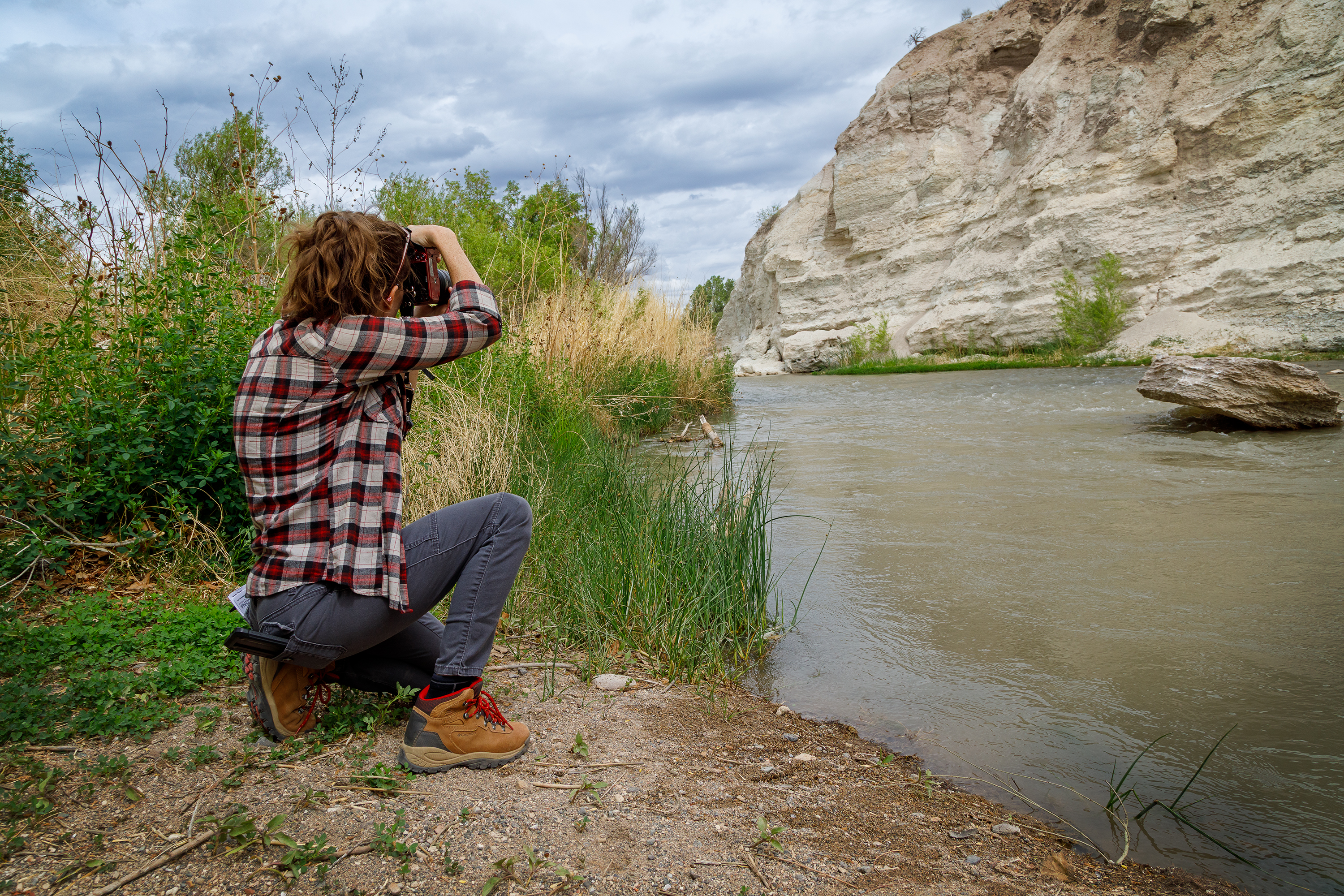 A person kneeling on the river bank taking a picture of the water flowing past white rock cliffs.
