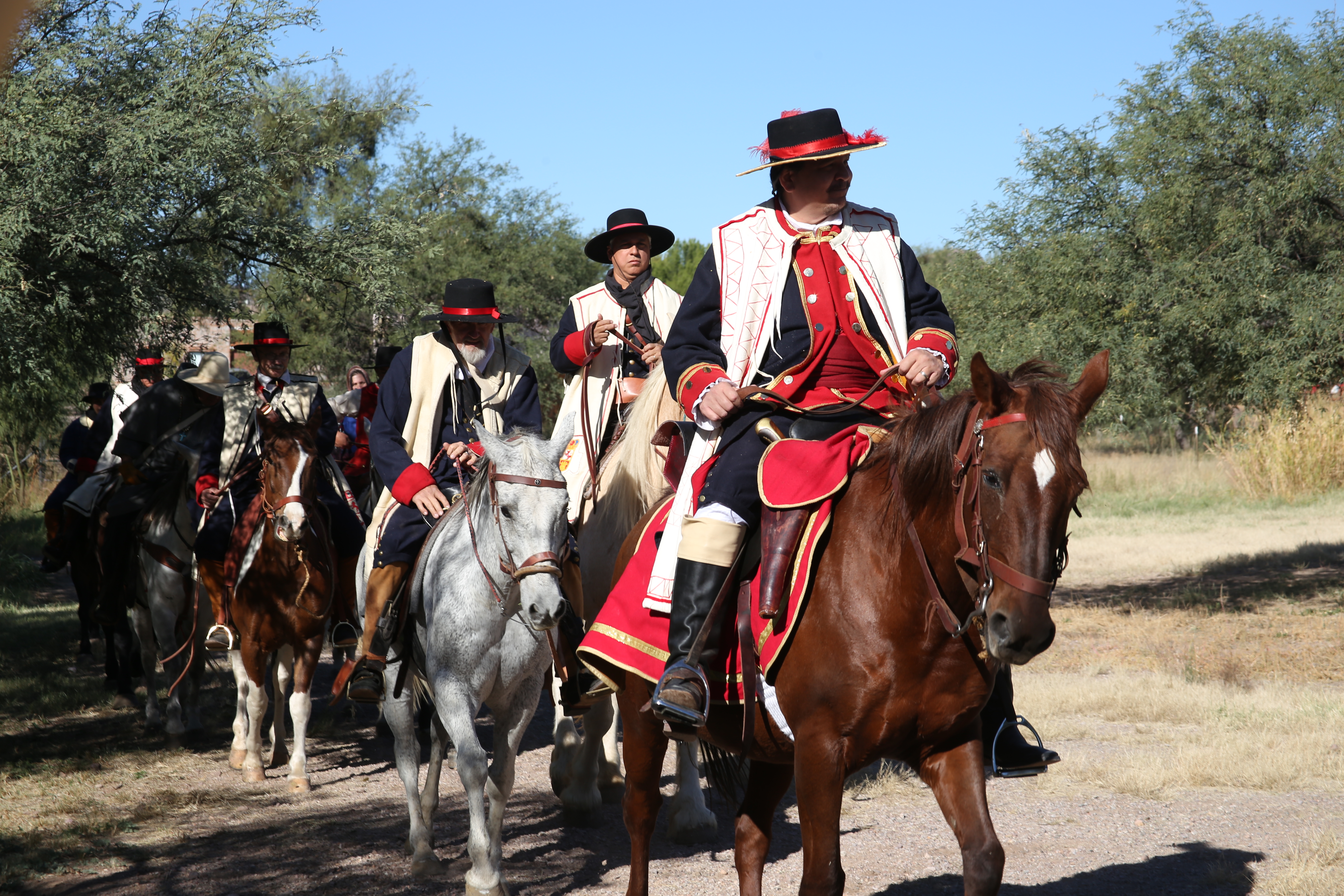 A procession of period reenactors dressed in Spanish military and settler costumes, riding on horseback through a mesquite-covered landscape.