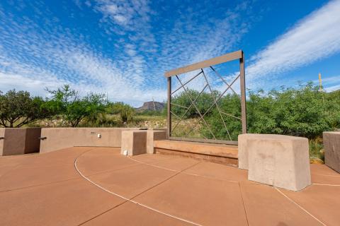 The stage of the amphitheater with a red concrete floor and a metal sculpture. The amphitheater is set among green trees and the sky is blue overhead.