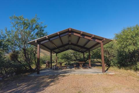 A shade ramada with picnic tables surrounded by green trees under a blue sky.