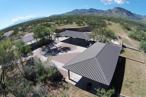 A group area with three large shade ramadas positioned in a U shape with a concrete and brick courtyard joining them. The ramada is set among green trees and mountains stand in the distance.