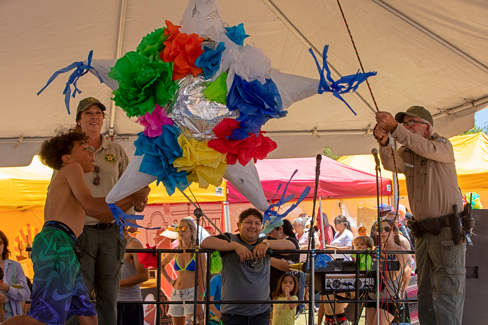 A child swings at a star-shaped multi-colored pinata, as two park rangers and a crowd looks on.