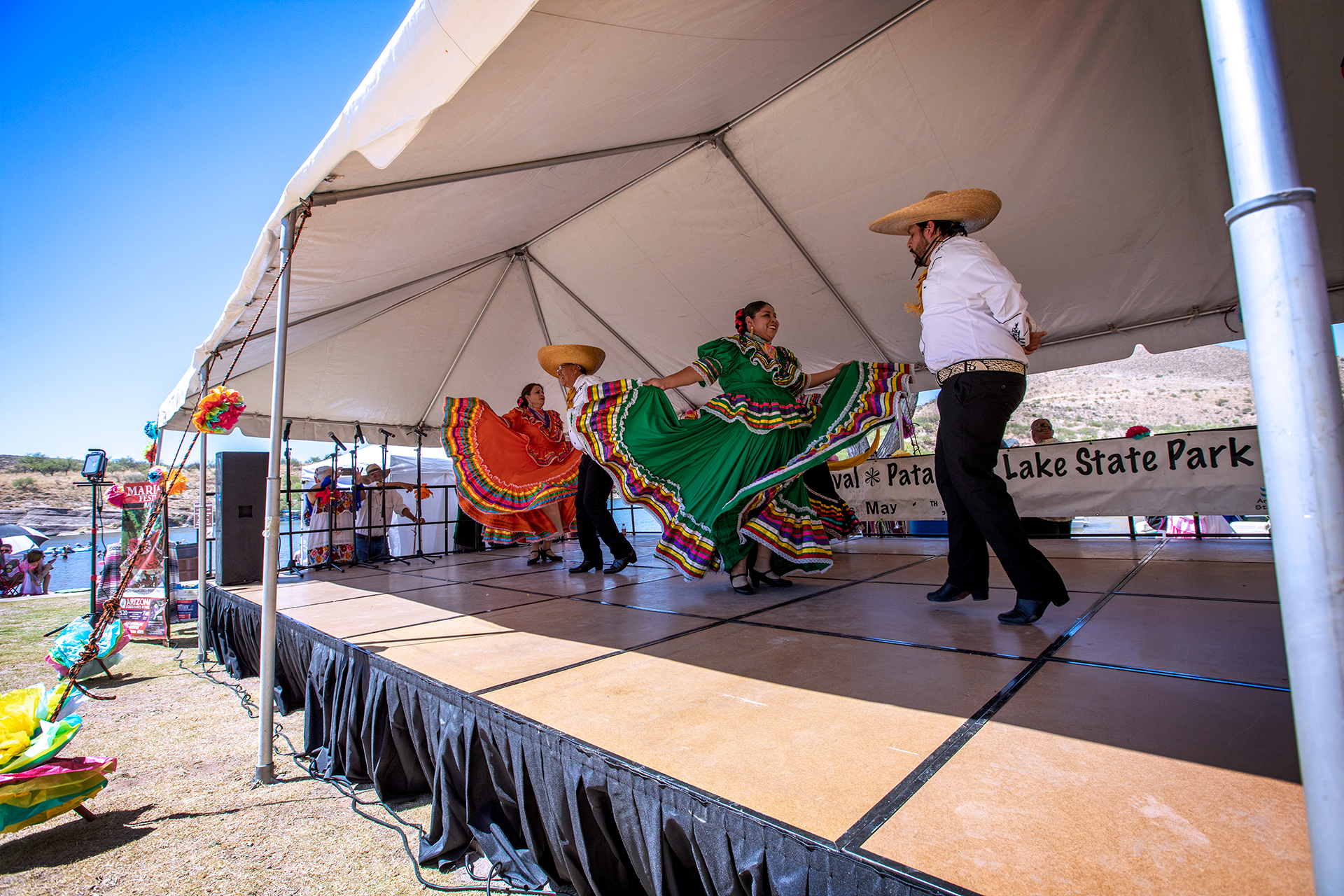 Mariachi dancers wearing colorful dresses and white shirts and sombreros dance on a stage.