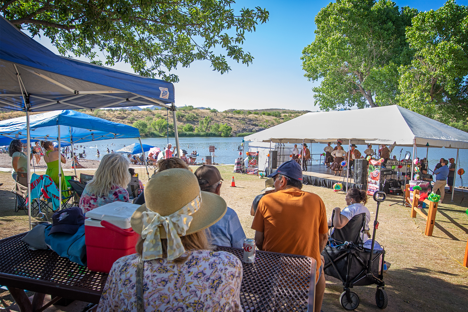A crowd sits under the shade of trees and popup tents, facing a stage that is set up in front of the lake.