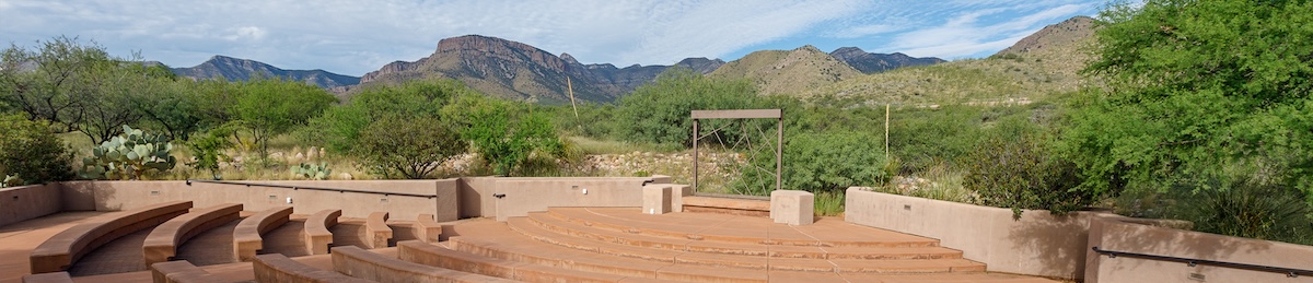 A photo of an amphitheater, with red concrete seats facing a raised stage in the front. The amphitheater is surrounded by mesquite trees and other desert vegetation under a blue sky partly covered with thin white clouds. The Whetstone mountains stand in t
