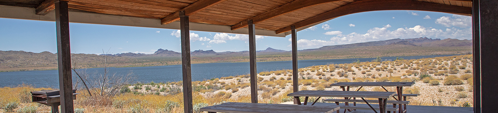 A view of Alamo Lake in the distance from a covered group ramada with picnic tables and a grill.