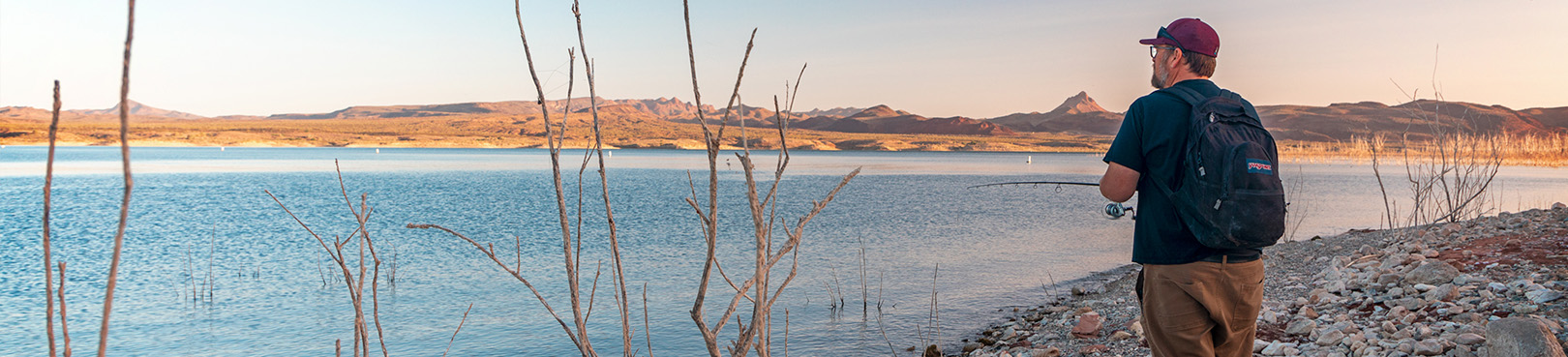 A person wearing a backpack fishing from the shore at Alamo Lake just after sunrise.