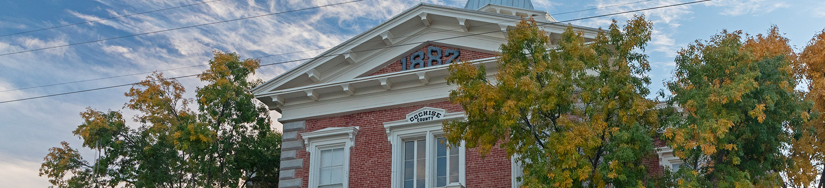 Looking up at the red brick courthouse framed by deciduous trees making their transition to fall colors on a partly cloudy day.