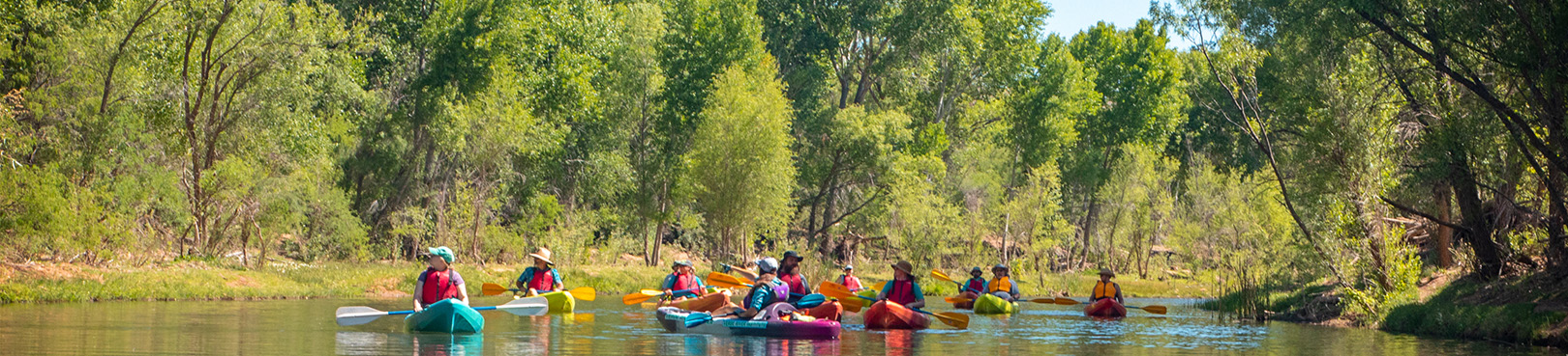 Kayakers float a relaxing section of the Verde River while surrounded by tall green riverside trees and partially covered with shadows.