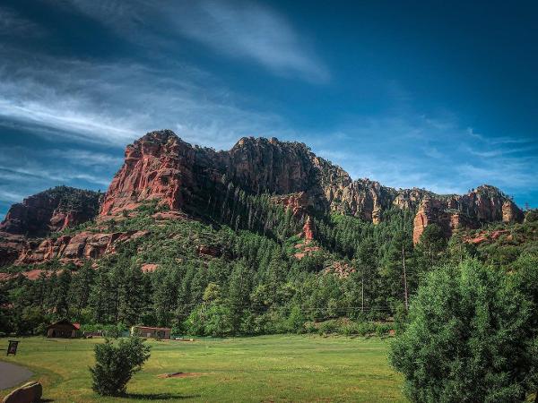 The massive red rocks covered in pine trees with green grass and a blue sky at Slide Rock State Park