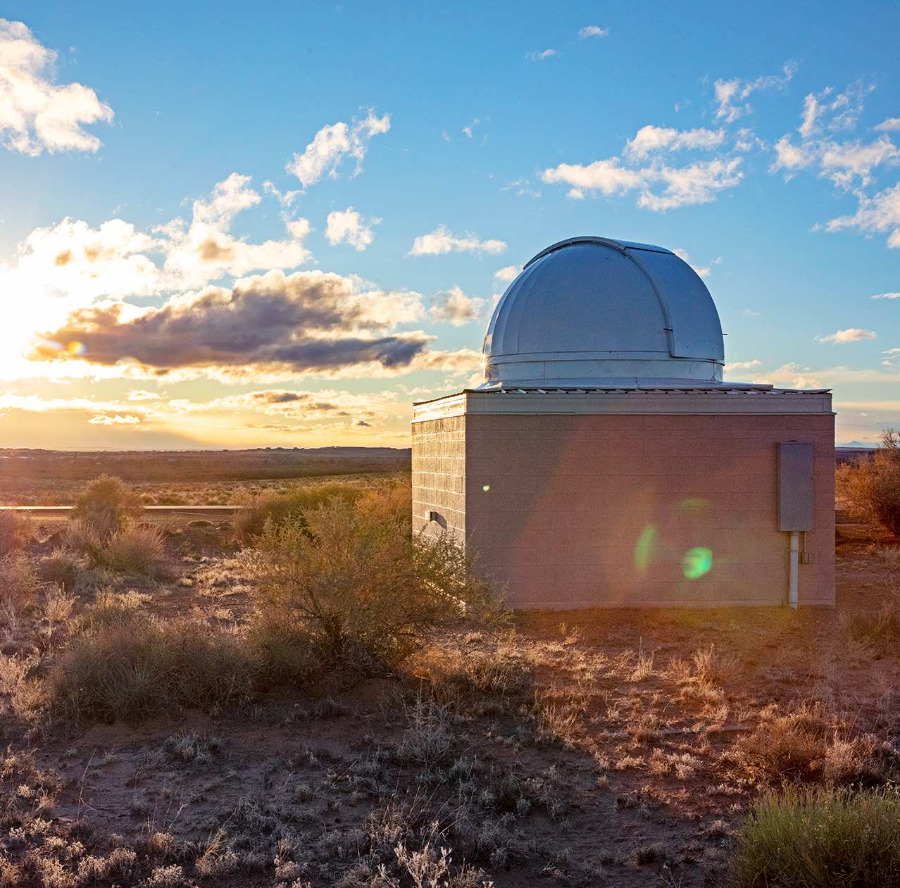 An observatory with a white domed roof sitting in a grassy landscape with the sun setting on the horizon.