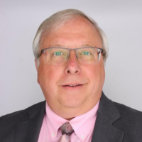 Headshot of Dr. Brent Bauer. He has white hair, wears glasses, and is wearing a black suitcoat, pink shirt, and a tie.