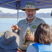 A photo of Louis Juers. He is holding a fake fish and talking to two children. A lake is visible in the background and a opoup tent overhead. He wears a tan hat and a state parks ranger shirt.